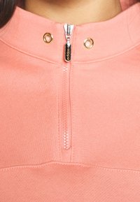 Suéter rosa claro con cuello alto, que cuenta con una cremallera frontal, ojales dorados y tejido texturizado. La cremallera tiene una lengüeta de metal negro con el nombre de la marca.