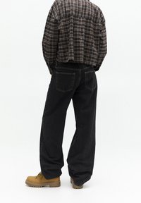 Jeans de mezclilla negra con un ajuste relajado, que cuentan con cinco bolsillos y costuras sutiles. Combinados con una camisa de franela a cuadros y botas de trabajo marrón.