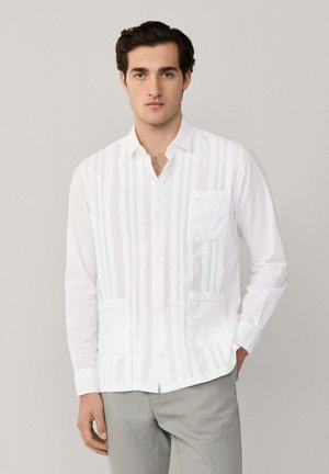 Jonge man met donker haar, gekleed in een wit overhemd met patroon en lichtgrijze broek, staand tegen een effen achtergrond.