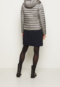 Femme portant une doudoune argentée, une jupe foncée, des collants noirs et des bottines noires, debout devant un fond clair uni.