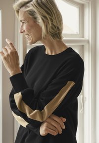 Black sweatshirt with long sleeves featuring beige stripes. Smooth fabric, relaxed fit, round neckline. Subject has short hair and is gesturing.