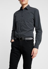 Chemise noire à boutons avec des pois blancs, à manches longues et col, associée à un jean noir et une ceinture grise.