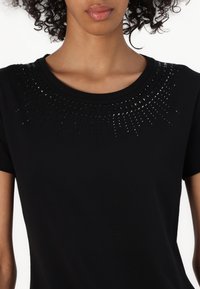 T-shirt nera con scollatura rotonda e maniche corte, decorata con un brillante motivo di borchie nere attorno al colletto.
