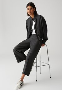 Grijze, gestructureerde jas met drukknoopsluiting en voorkantzak; bijpassende grijze wijde broek; witte shirt; wit met zwarte sneakers; zittend op een kruk.