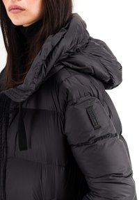 Veste en duvet noire avec une grande capuche, un tissu texturé et un patch logo visible sur la manche. Fermeture à glissière et détails avec cordon de serrage.