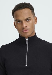 Juodas zip-up megztinis su aukšta apykakle, pagamintas iš tekstūruotos mezginio medžiagos, su sidabriniu užtrauktuku ir minimalistiniu dizainu.