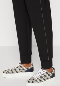 Beige rakstainie sporta apavi ar tumši zilu akcentu, baltu zoli un auklām; apvienoti ar melnajiem pieguļošajiem sporta biksēm ar sānu šuvēm.