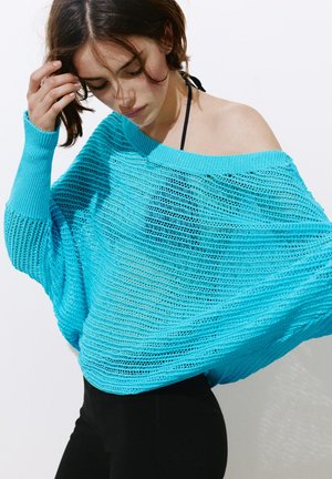 Jeune femme ajustant ses cheveux, vêtue d’un pull tricoté turquoise ample à épaules dénudées, porté sur un pantalon noir et un top noir à col halter.