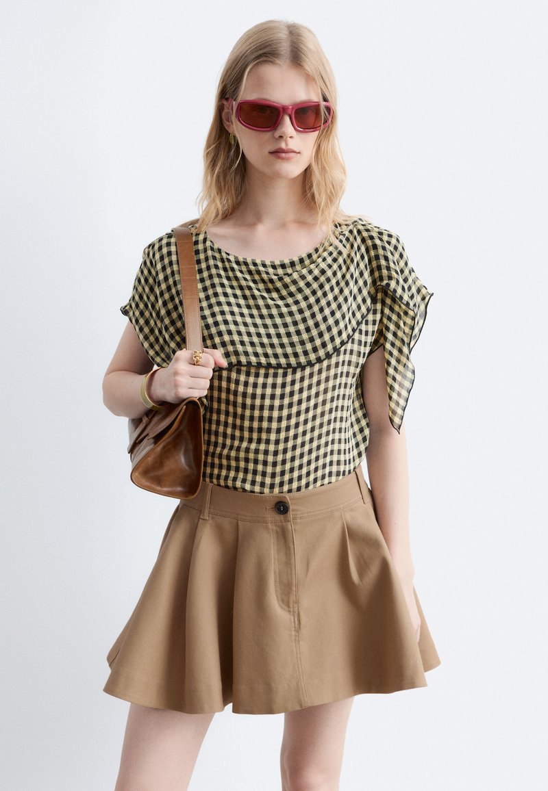 Jeune femme portant des lunettes de soleil rouges, un haut à carreaux noir et beige, une jupe beige et tenant un sac à bandoulière marron.
