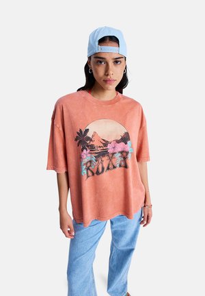T-shirt oversize rose-orangée avec un col rond et des manches courtes. Présente un imprimé graphique de montagnes et de fleurs. Portée avec un jean bleu clair.