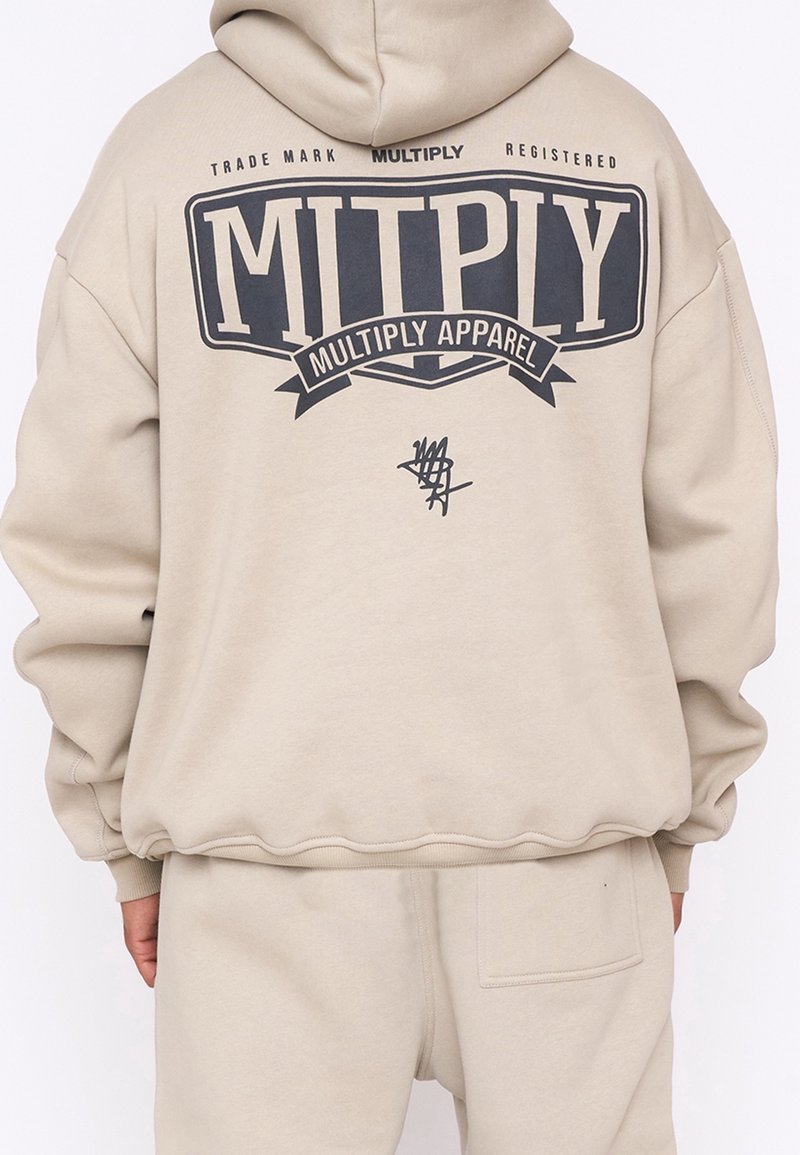 Osoba v béžové mikině a kalhotách, mikina vzadu s potiskem "MULTIPLY" a "Multiply Apparel" tučnými černými písmeny.