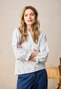 Femme aux longs cheveux blonds portant un chemisier blanc à motifs floraux et un jean bleu, se tenant les bras croisés devant un mur beige uni.