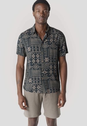 Hombre vistiendo una camisa de manga corta con botones y patrones geométricos, y pantalones cortos beige texturizados, de pie frente a un fondo claro y liso.