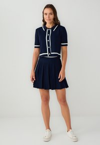 Navy gestricktes Zweiteiler-Outfit mit plissiertem Rock und kurzärmligem Oberteil mit weißem Besatz und Knöpfen, kombiniert mit weißen Sneakers.