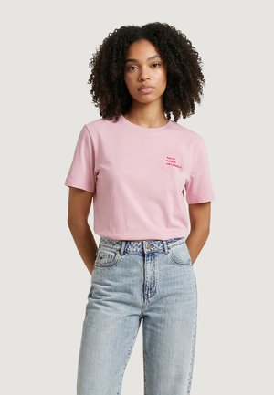 Giovane donna con capelli ricci che indossa una maglietta rosa chiaro con testo rosso e jeans blu chiaro a vita alta, in piedi con le mani dietro la schiena.