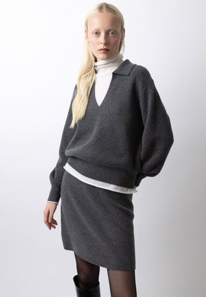 Donna bionda che indossa un maglione a coste grigio scuro con maniche a palloncino e gonna abbinata sopra una maglia a collo alto bianca, in piedi.