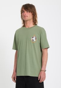 Jeune homme aux cheveux mi-longs portant un t-shirt vert olive avec un motif de globe sur la poitrine et un pantalon noir sur fond blanc.