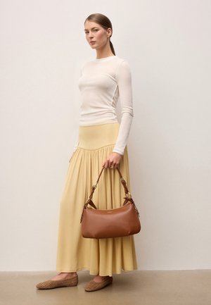 Vrouw met een wit shirt met lange mouwen, een gele maxirok en gevlochten platte schoenen, die een bruine leren handtas met korte hengsels vasthoudt.