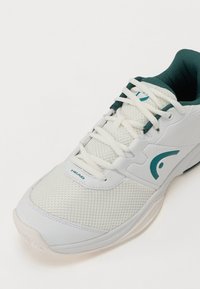 Hvid atletisk sko med mesh og syntetisk overdel, teal accenter og en tekstureret sål. Har snørebånd og et logo på siden.