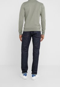 Sudadera gris con puños y dobladillo acanalados, combinada con jeans azul oscuro. Zapatos blancos con detalles azules. Vista desde atrás.