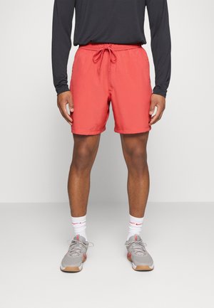 Mann trägt rote Kordelzugshorts, graue Sneakers, weiße Kniestrümpfe und ein langärmeliges schwarzes Shirt und steht vor einem einfarbigen Hintergrund.