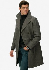 Grijs herringbone wol overjas met grote revers, dubbelgebreid ontwerp, zijzakken en een zachte textuur, gedragen over een donkere trui.
