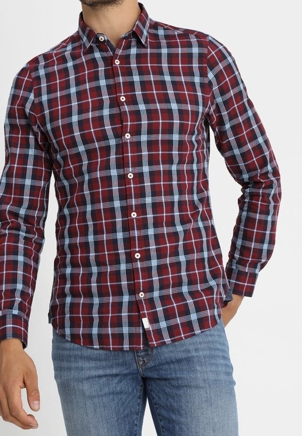 Homme portant une chemise à carreaux rouge et bleu à manches longues boutonnée, avec un jean bleu clair, debout devant un fond blanc.