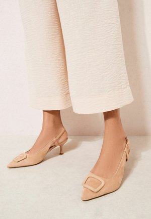 Picioare cu sandale cu toc mic tip kiten, din piele întoarsă bej, cu catarame decorative mari, asortate cu pantaloni largi crem, texturati, tăiați deasupra gleznelor.