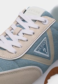 Close-up af en blå og beige Guess-sneaker med hvide snørebånd og en trekantet Guess-logo på siden.