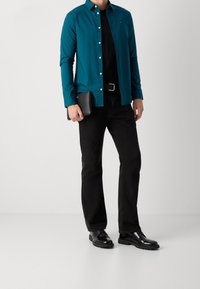 Turkooise overhemd over een zwart T-shirt, gecombineerd met zwarte jeans en glanzende zwarte schoenen. Houdt een zwarte portemonnee in de hand.