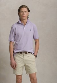 Ανοιχτό μοβ πόλο μπλουζάκι με γιακά και κοντά μανίκια, σε συνδυασμό με μπεζ σορτς. Η μπλούζα έχει διακριτά σχέδια και ένα λογότυπο στο στήθος.