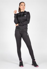 Zwarte sportjack met ritssluiting en capuchon, gecombineerd met aansluitende zwarte legging. Beide hebben een subtiel logo en contrasterende roze accenten op de schoenen.