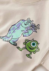 Felpa beige con una stampa grafica di due mostri dei cartoni animati. Uno ha macchie blu, l'altro è verde con un occhio. Design casual.