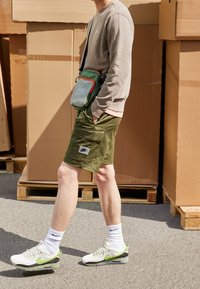 Olivgrüne Shorts mit mehreren Taschen, kombiniert mit einem hellbraunen Sweatshirt und weißen Sneakers mit grünen Akzenten. Eine grüne Umhängetasche wird getragen.