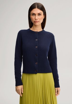 Femme aux cheveux raides et foncés porte un cardigan bleu marine boutonné et une jupe plissée jaune-vert sur un fond clair uni.
