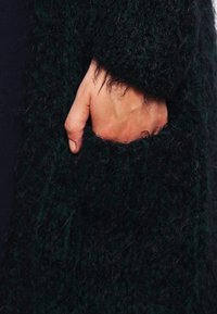 Mörkgrön texturerad cardigan med en stor, fluffig stickad design och en framficka. Tyget ser mjukt och varmt ut.