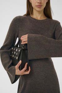 Musta käsilaukku, jossa on metalliset pyöreät niitit, jota pitää henkilö, joka on pukeutunut pitkään, ribattyyn, ruskeaan neuleeseen, jossa on levenevät hihat.