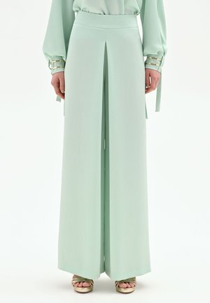 Donna che indossa pantaloni verde menta a gamba larga con piega frontale, abbinati a una camicetta a maniche lunghe coordinata e tacchi aperti dorati.