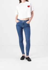 Camiseta blanca recortada con un gráfico de un corazón rojo, combinada con jeans ajustados azules y mocasines negros. Diseño de atuendo simple y casual.