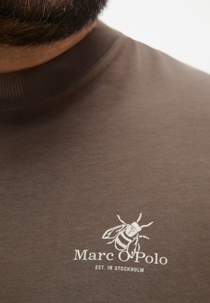 Nahaufnahme eines braunen Marc O'Polo Hemds mit weißem Bienen-Logo und dem Text "EST. IN STOCKHOLM" auf der Brust in Kragenhöhe.