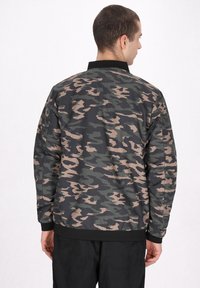 Camouflagejakke i sort, grøn og beige; letvægtsmateriale, ribbet krave og manchetter, samt en afslappet pasform. Bagsiden vises.