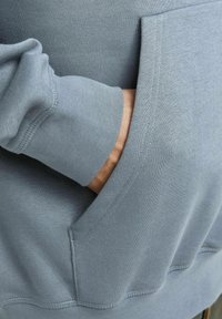 Sudadera de color azul claro con una textura suave, que cuenta con un bolsillo tipo canguro y puños acanalados. Se pueden ver las costuras a lo largo de los bordes.