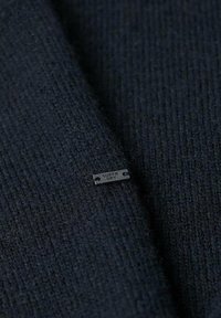 Tejido de punto negro con un patrón texturizado, que presenta una pequeña etiqueta de metal etiquetada como "SUPER DRY" cosida en la trama.