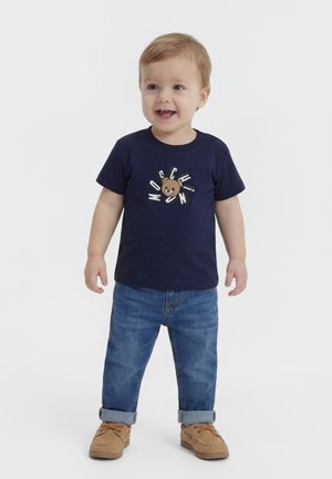 Νηπιακό παιδί που φοράει μπλε μπλουζάκι με γραφικό αρκούδας και τη λεζάντα "Moschino", μπλε τζιν παντελόνια με ρεβέρ και ταμπά δετά παπούτσια.