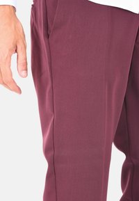 Pantaloni borgogna su misura realizzati in tessuto testurizzato, con taglio dritto e tasche laterali. Una mano visibile riposante vicino alla vita.