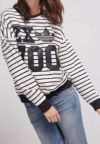 Pruhovaná černobílá mikina se silným potiskem loga, žebrovanými manžetami a lemem. V kombinaci se světle modrými džíny. Uvolněný design.