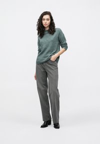 Sive široke kavbojke z volnenim puloverjem v turkizni barvi, ki ima okrogli izrez in dolge rokave, na enobarvni beli podlagi.