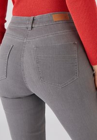 Jeans grises de mezclilla con un ajuste ceñido, que cuentan con dos bolsillos traseros y una etiqueta de cuero marrón en la cintura. Superficie texturizada.