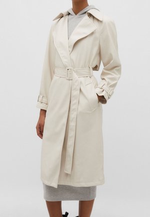 Personne portant un trench-coat beige ceinturé par-dessus une robe à capuche grise, debout avec une main dans la poche du manteau devant un fond uni.