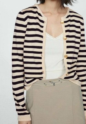 Person iført en sort og beige stribet knapcardigan over en hvid top og en beige nederdel med højtaljede og dobbelt spænde bælte.